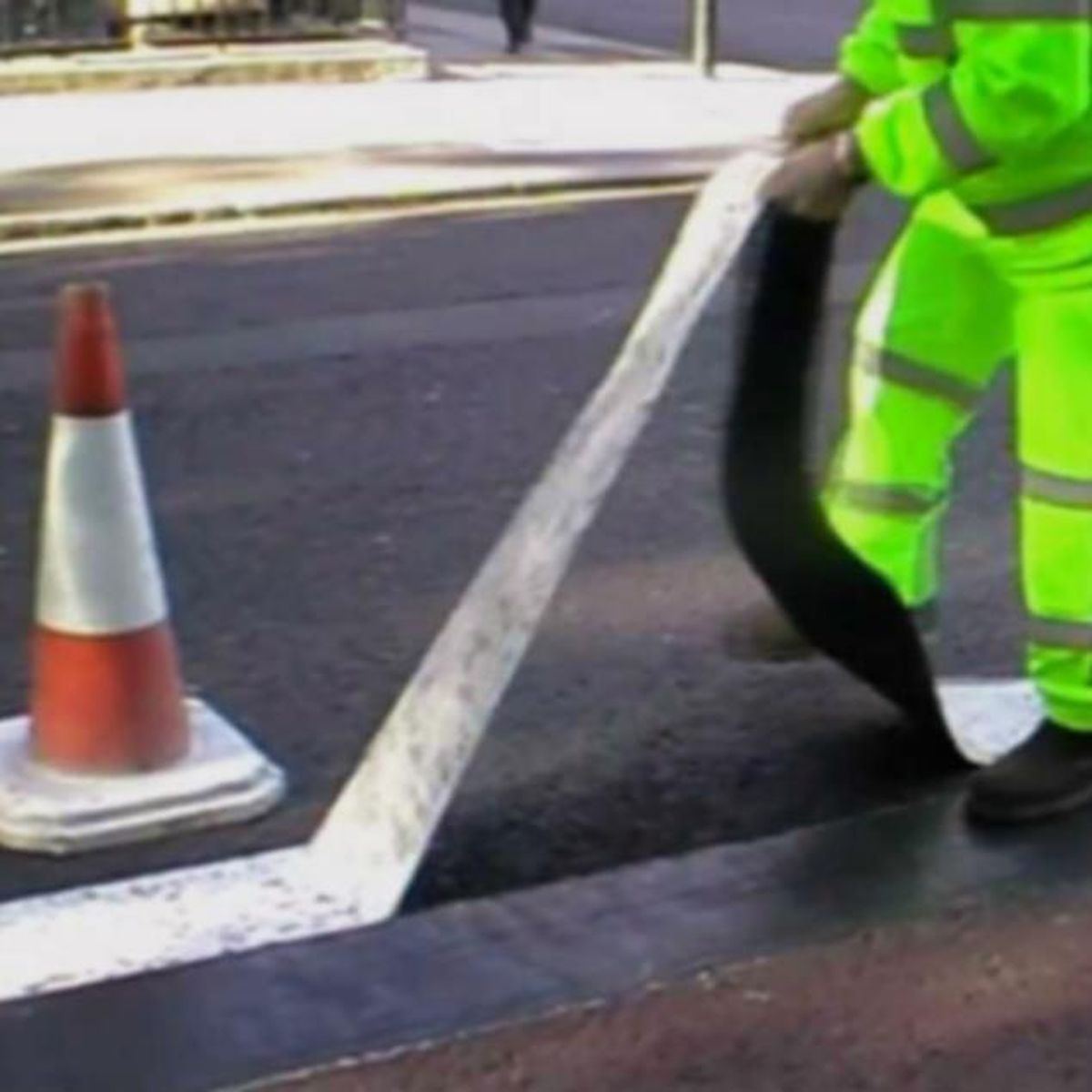 Operative in high-visibility clothing applying temporary road marking paint beside a traffic cone